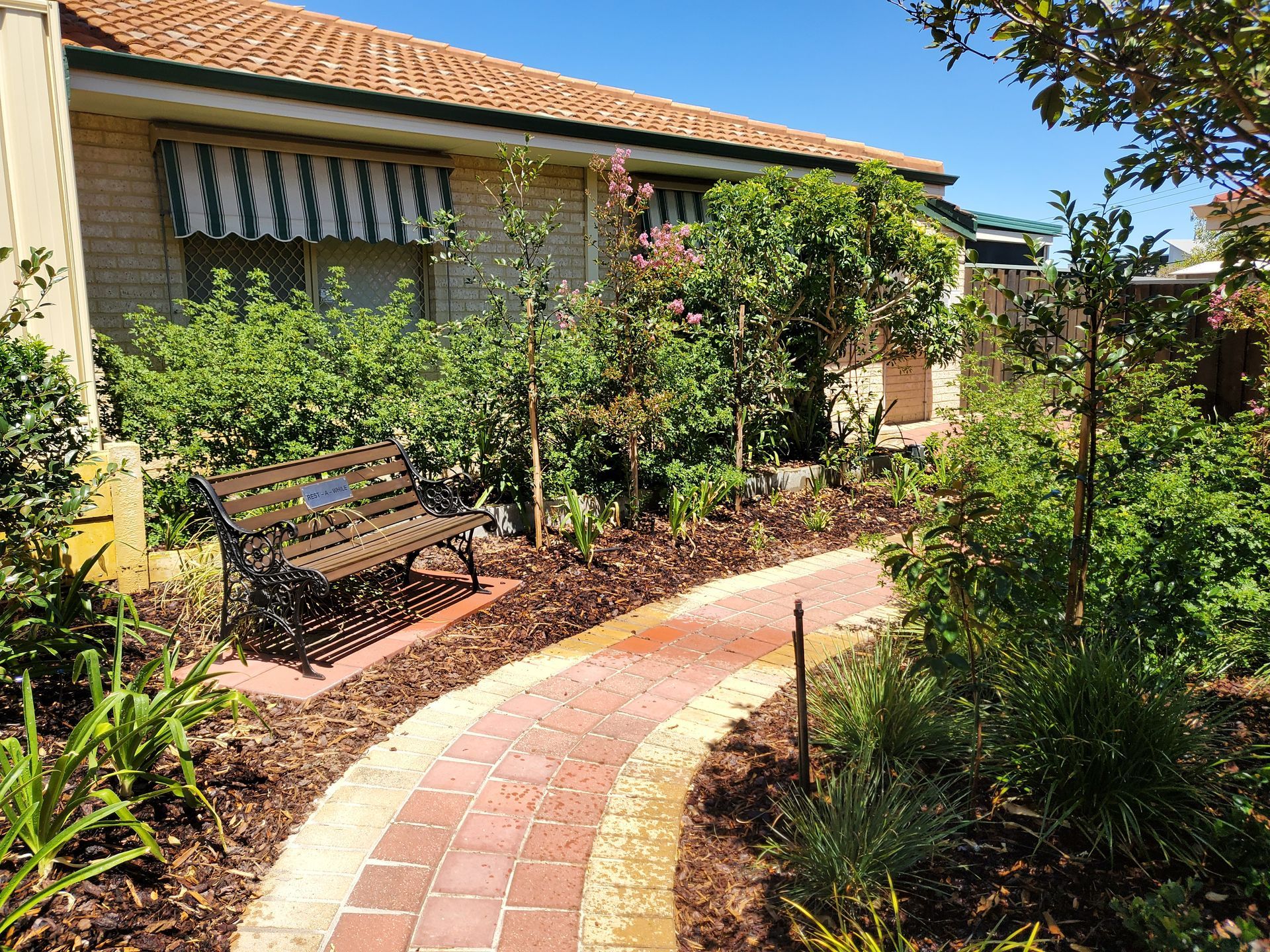 Brick path winds through a garden towards a building with striped awning. Bench sits alongside.