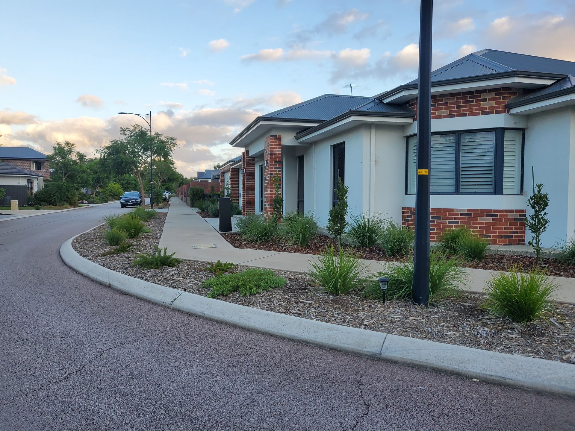 Curving residential street with light-colored houses and landscaping under a cloudy sky.