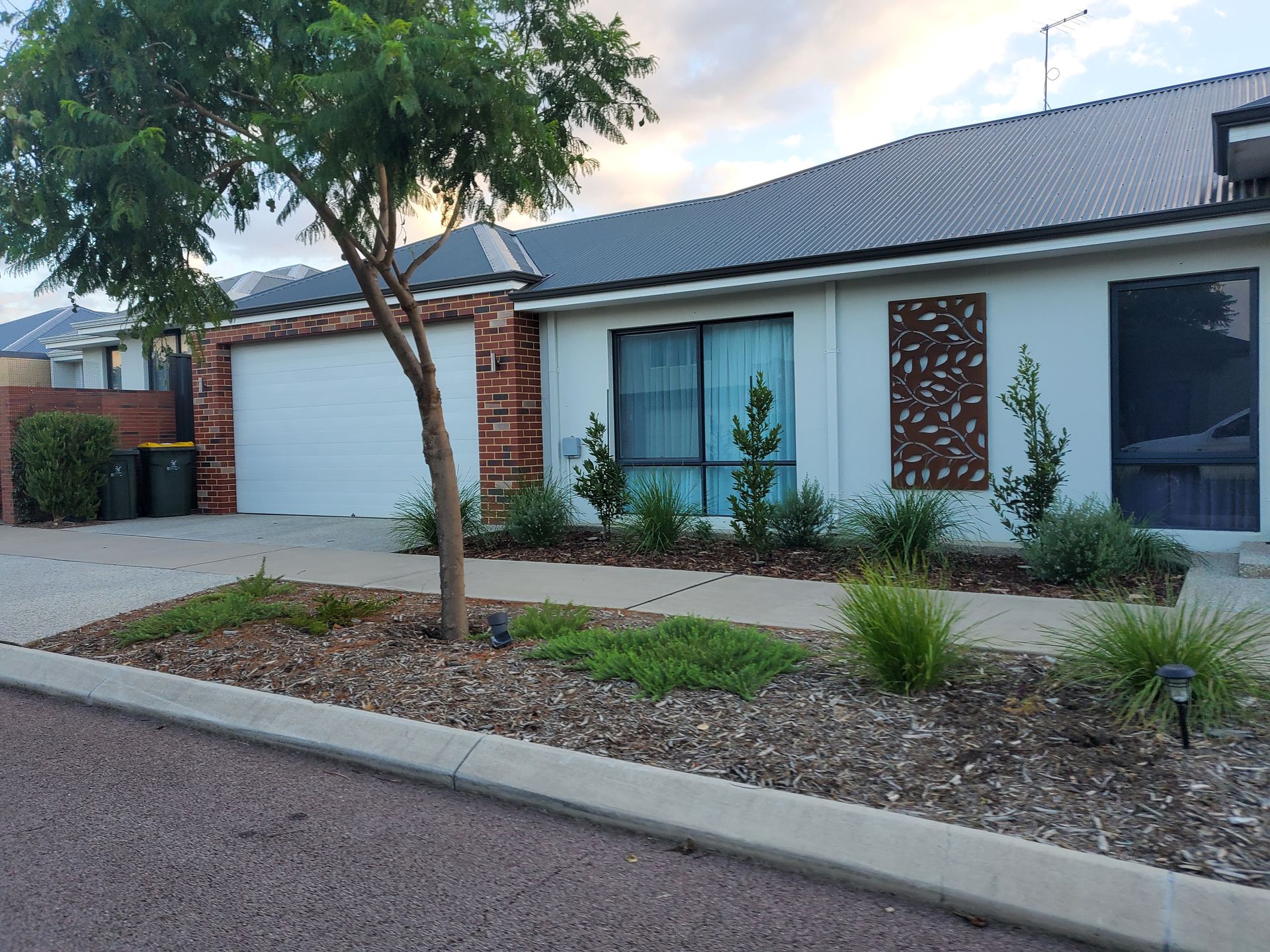 A white house with a garage, front yard with landscaping, and a tree on a paved street.