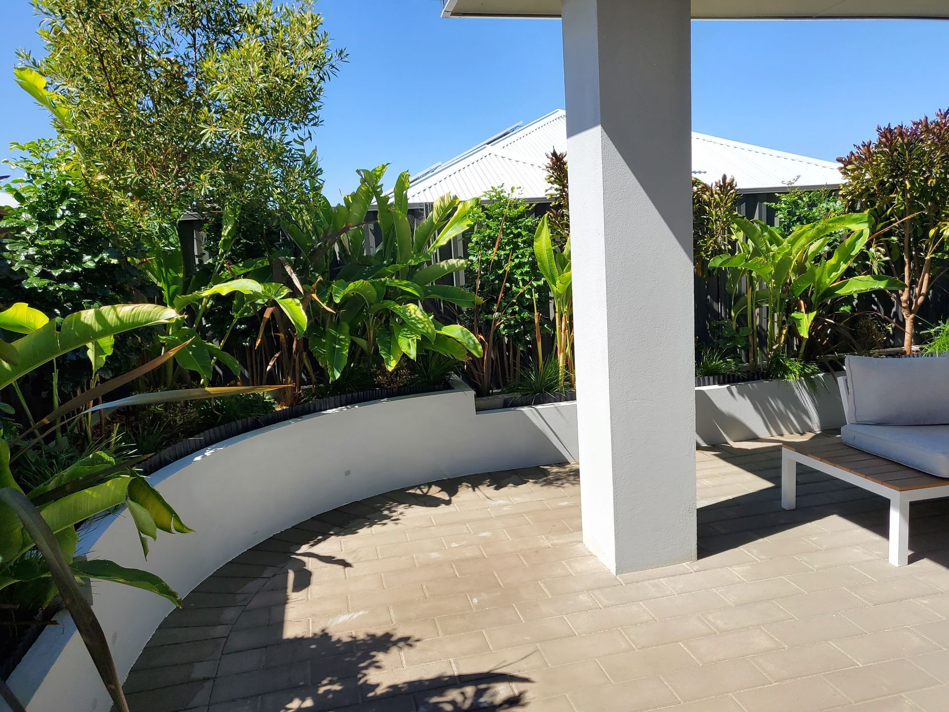 Outdoor patio with a white pillar, curved wall, lush plants, and a house in the background.