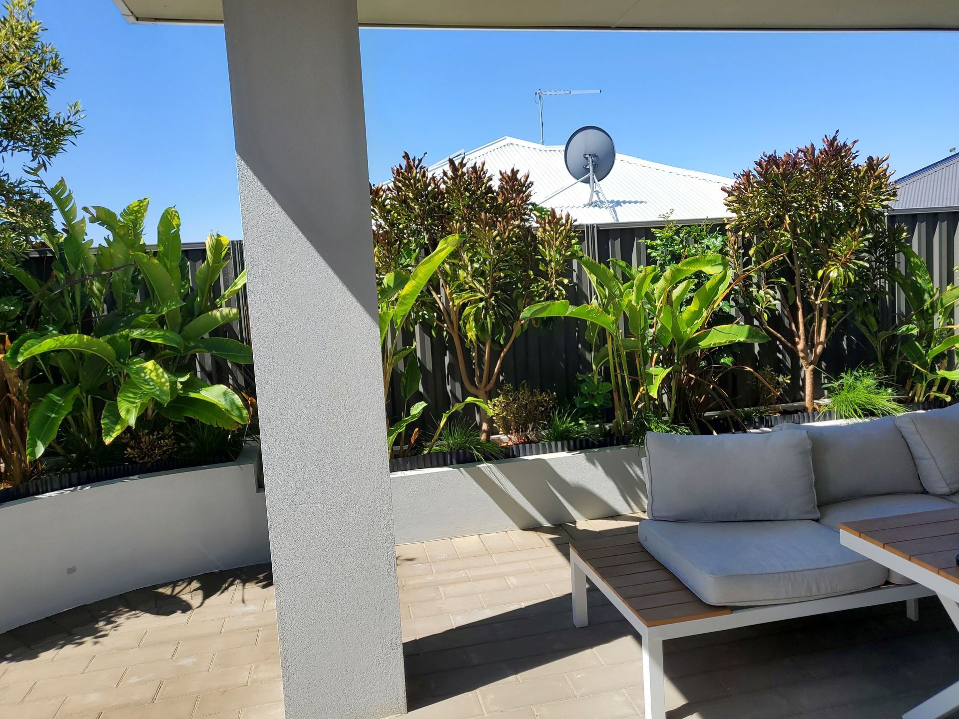 Patio with white furniture, lush green plants, and a house with a satellite dish on a sunny day.