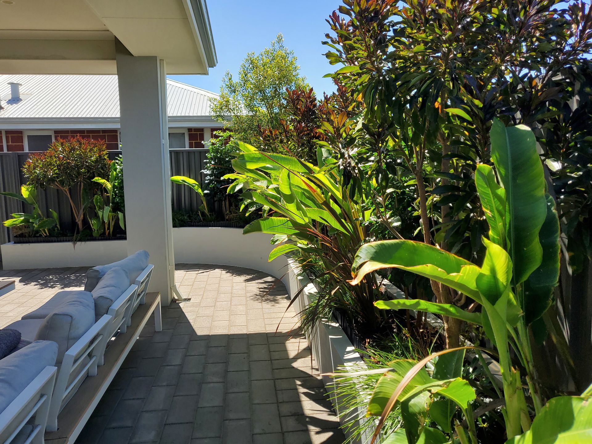 Patio with white couches, brick walkway, and lush green plants. Sunny day, outdoor setting.