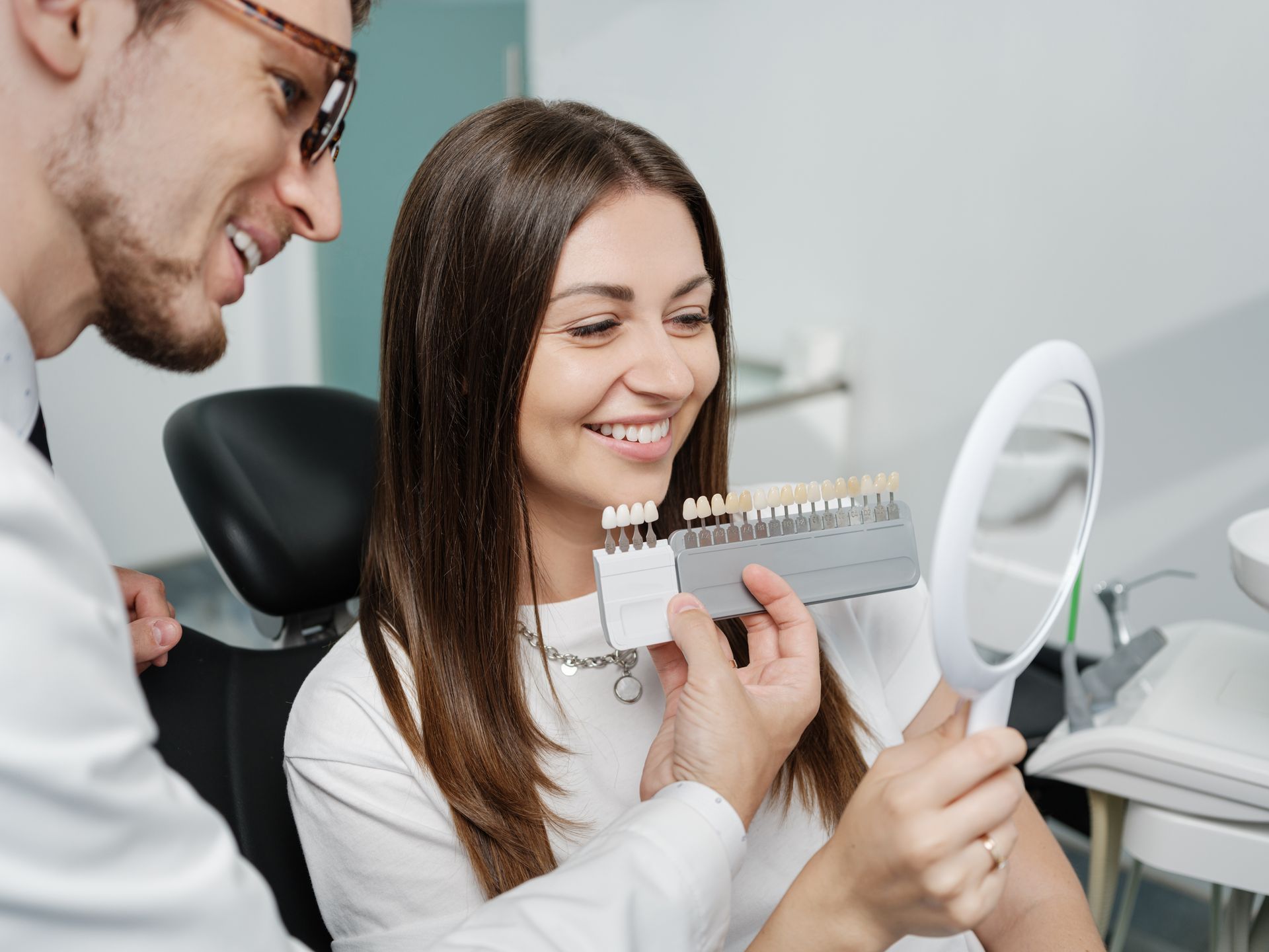 Patient in dentist's seat while doctor choosing the shade of dental veneer according to color chart Young man dentist in white coat show veneers enamel color palette for beautiful patient woman.
