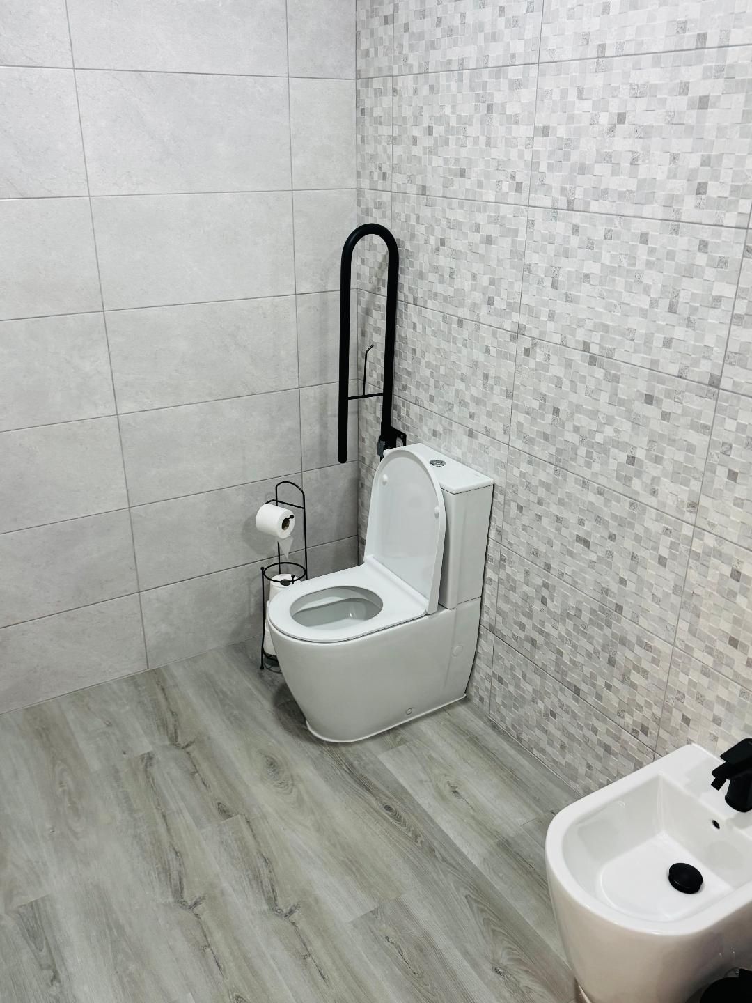 A white toilet with a black fold-up grab bar and a nearby toilet paper stand in a bathroom with light-toned tiled walls.