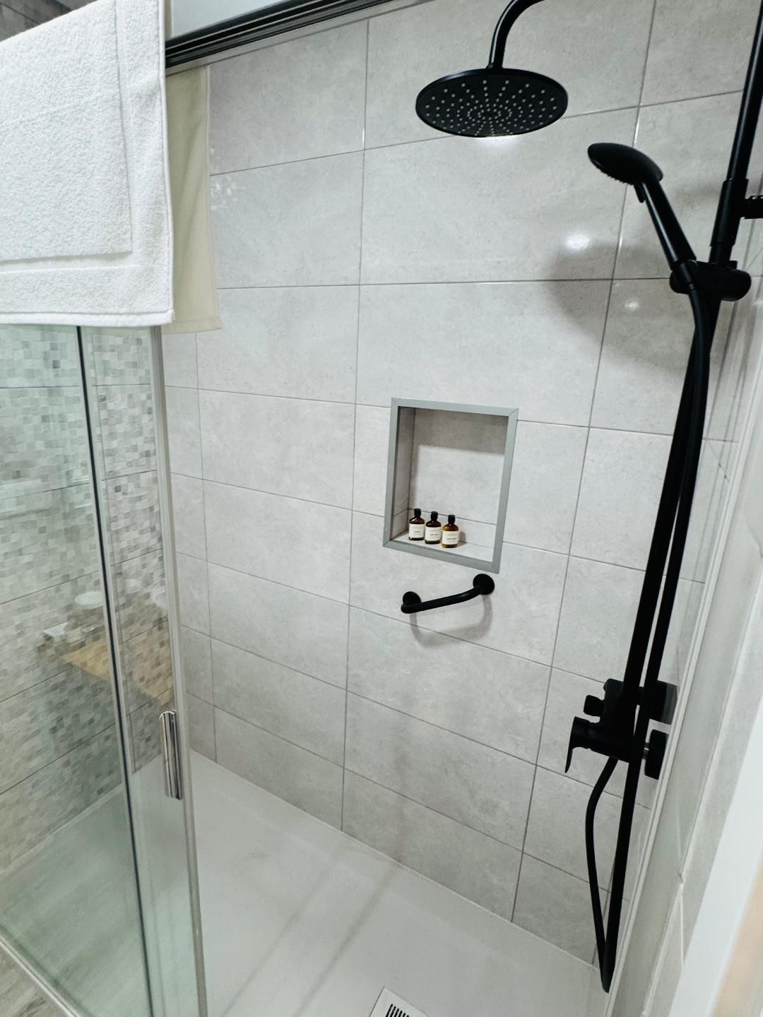 A modern glass-enclosed shower with gray tiled walls, a black rainfall showerhead, handheld sprayer, and a storage niche.