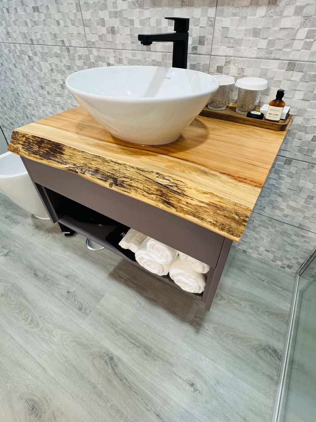 A modern bathroom vanity with a live-edge wood countertop, white vessel sink, black faucet, and open storage for towels.