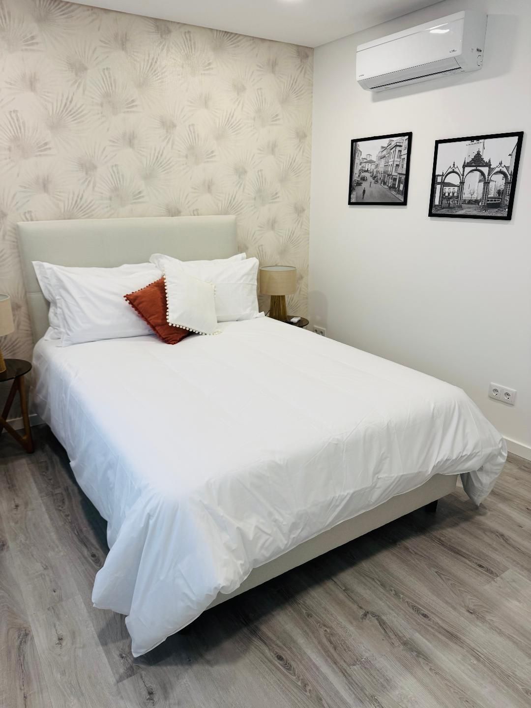 A modern bedroom with a queen bed featuring white bedding, a rust-colored accent pillow, floral wallpaper, and art prints.