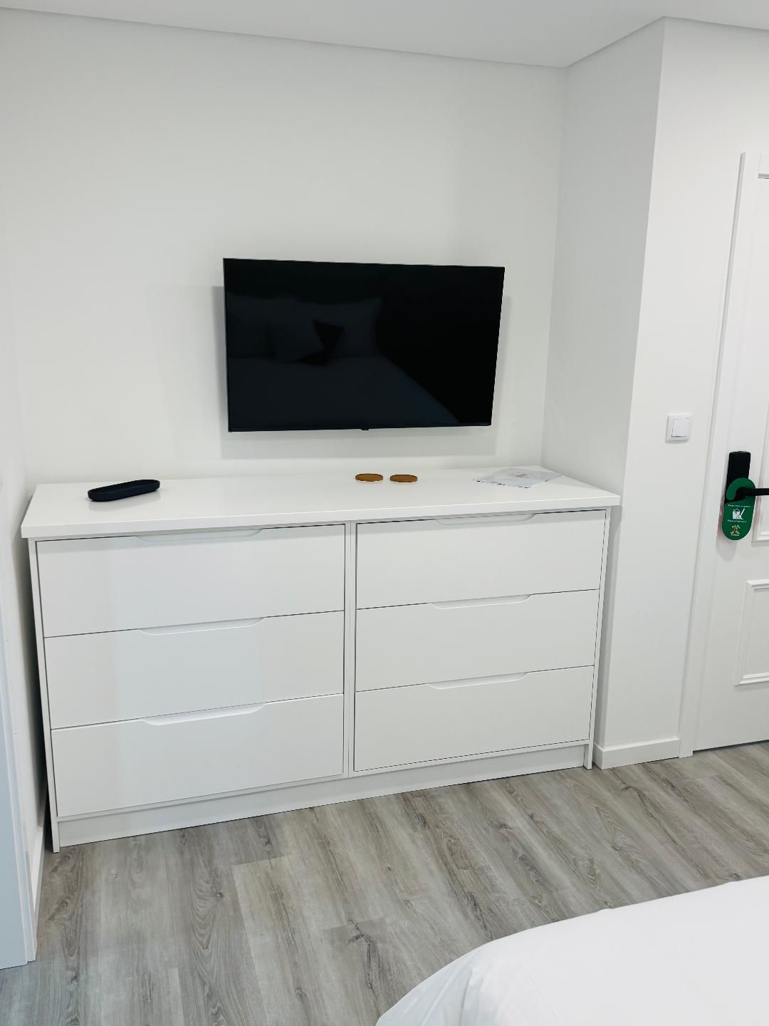 A wall-mounted TV sits above a white, six-drawer dresser in a room with light wood-look flooring.