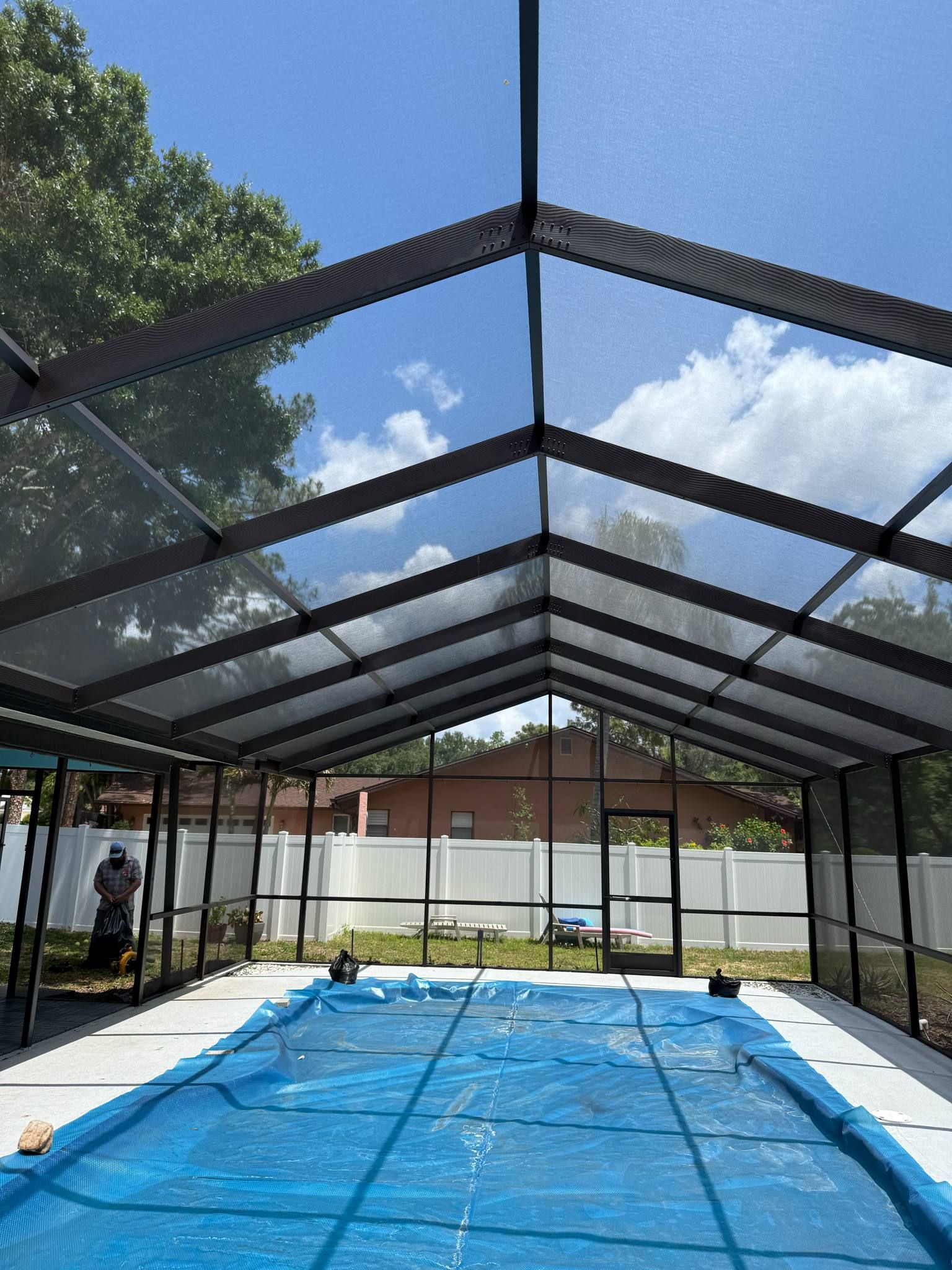 A blue pool cover lies in an enclosed pool area featuring a metal-framed screen roof under a bright, sunny sky.