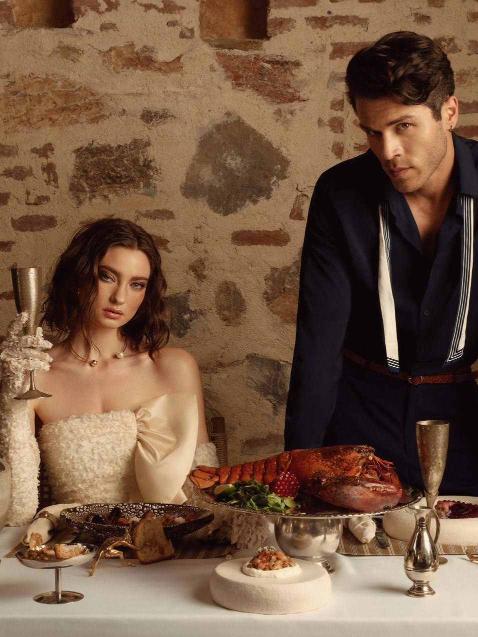 A person in a formal cream gown and gloves holds a glass across from a person in a dark shirt at a table with a roast.
