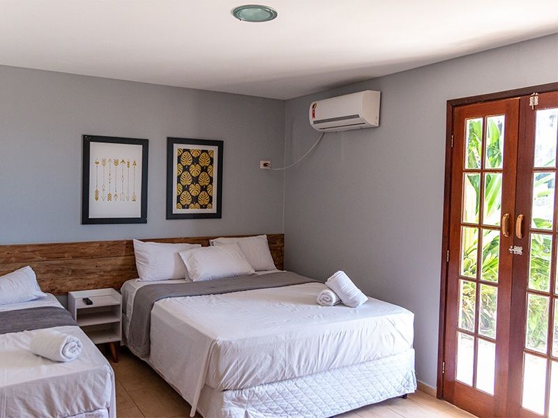 Quarto com duas camas, paredes cinza, cabeceira de madeira, ar condicionado e portas francesas.