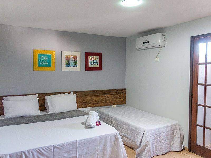Quarto com cama de casal, cama de solteiro e obras de arte na parede. Ar-condicionado acima da cama de solteiro. Porta à direita.