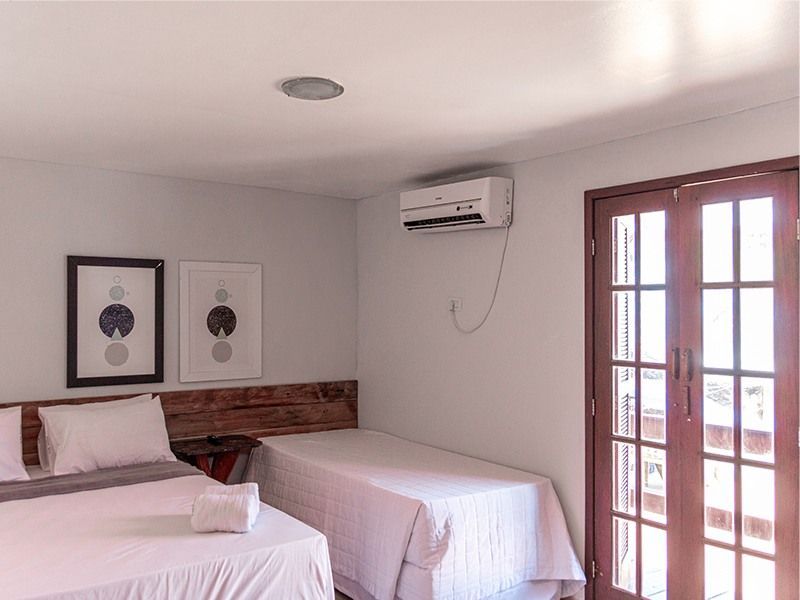 Quarto com duas camas, lençóis brancos, cabeceira de madeira e portas para varanda. Duas obras de arte emolduradas.