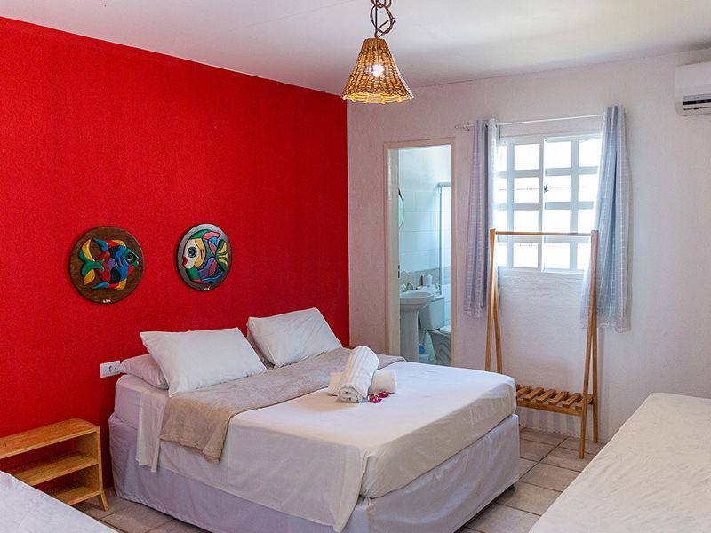 Quarto com parede de destaque em vermelho, cama de casal, obras de arte e banheiro privativo.