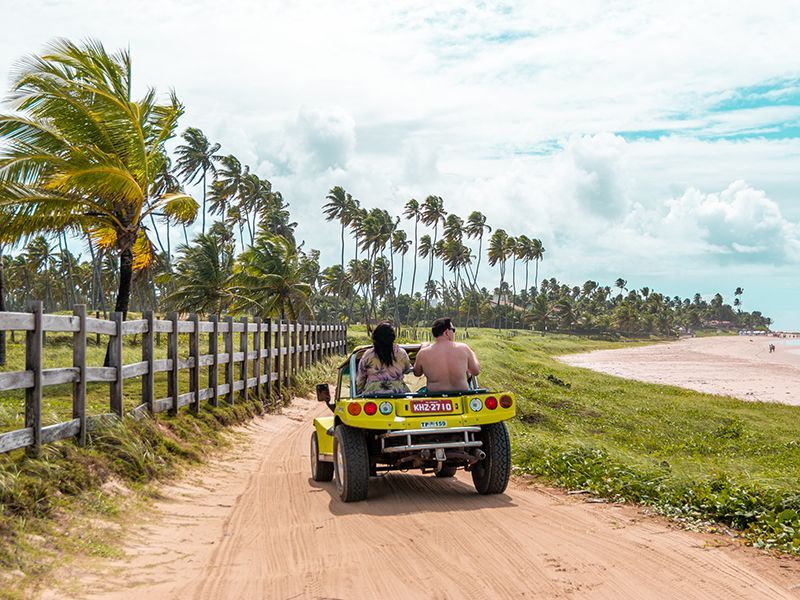 Um buggy amarelo de praia trafegando em uma estrada de areia ao lado de uma praia com palmeiras e uma cerca de madeira.