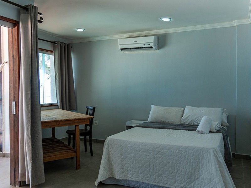 Quarto com cama de casal, mesa, cadeira e ar condicionado de parede. Paredes azul-claras, roupa de cama clara.