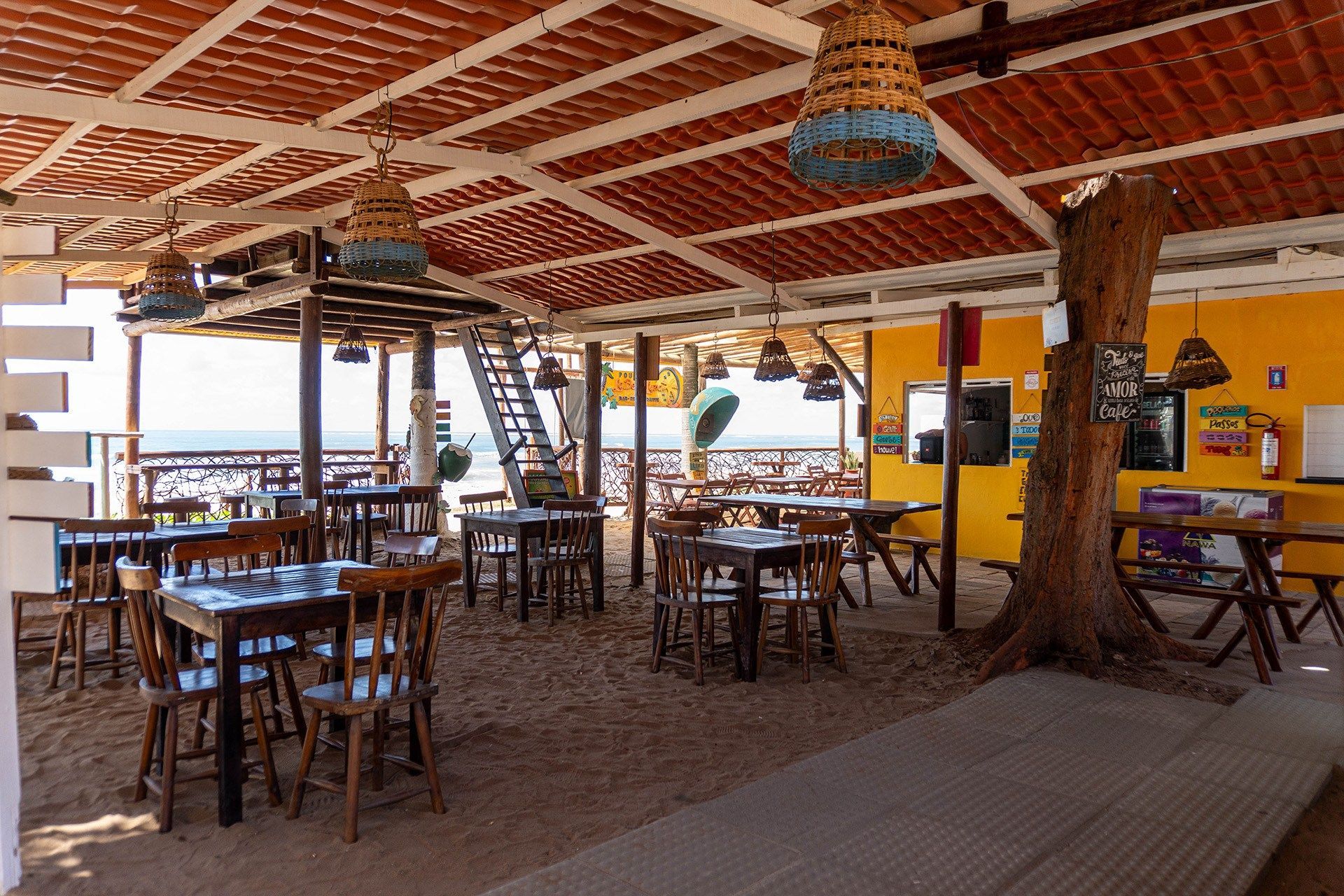 Restaurante à beira-mar com mesas e cadeiras sob um toldo; oceano visível ao fundo.