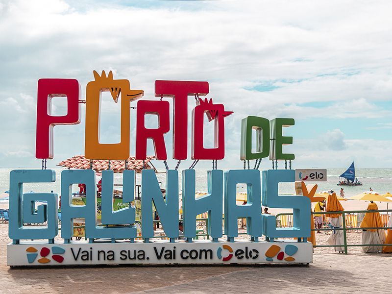 Placa colorida com a inscrição "Porto de Galinhas" em uma praia, sob um céu nublado.