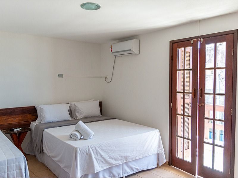 Quarto com cama de casal, cama de solteiro pequena e porta para uma varanda. Paredes brancas, acabamento em madeira.