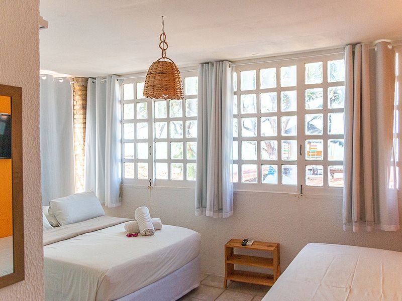 Quarto com duas camas, roupa de cama branca, janelas amplas, luminária de vime e mesa de cabeceira de madeira.