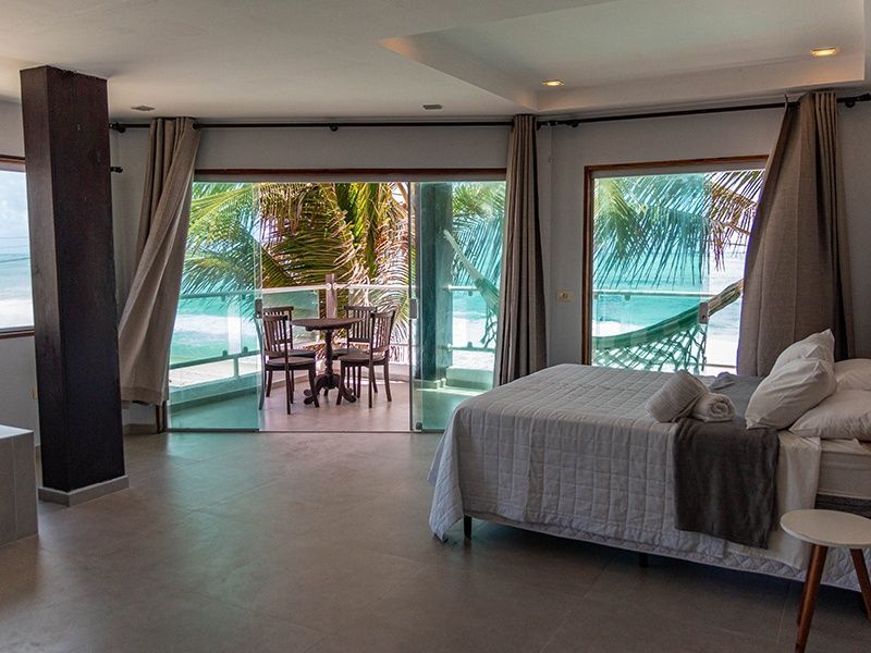 Quarto com vista para o mar; cama, mesa, cadeiras e rede na varanda.