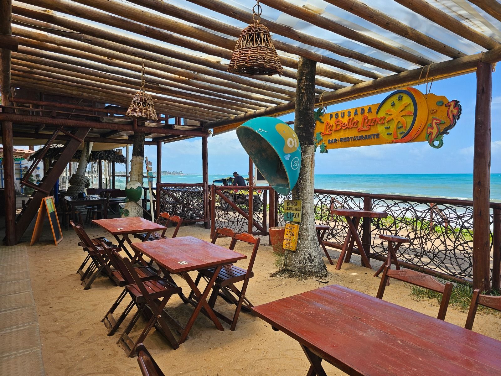 Restaurante ao ar livre com vista para o oceano, mesas de madeira e uma placa peculiar.