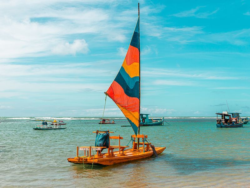 Veleiro de madeira com vela colorida no oceano, outros barcos ao longe, dia ensolarado.