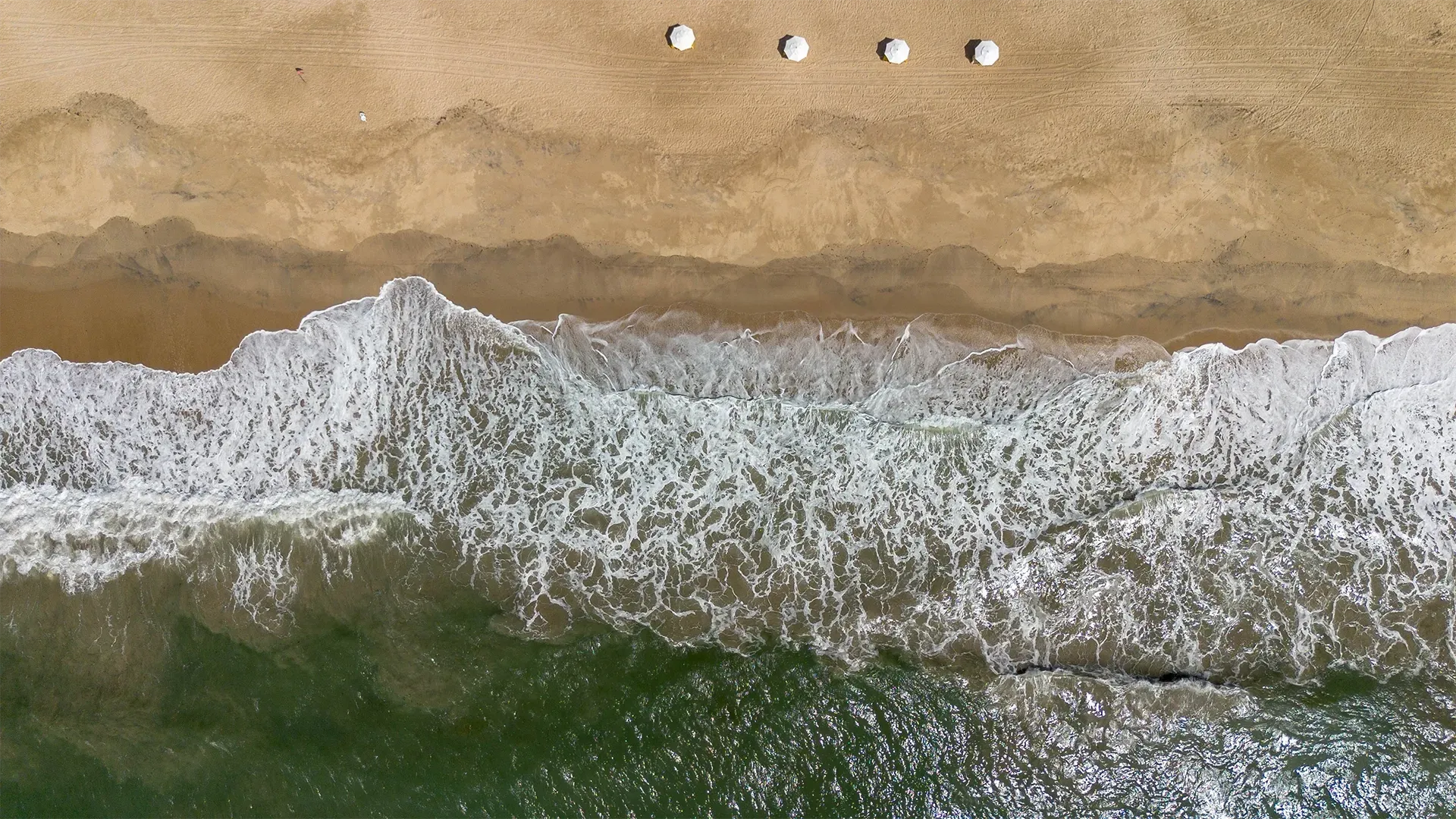 Vista aérea das ondas quebrando em uma praia de areia. Três objetos brancos estão na areia.