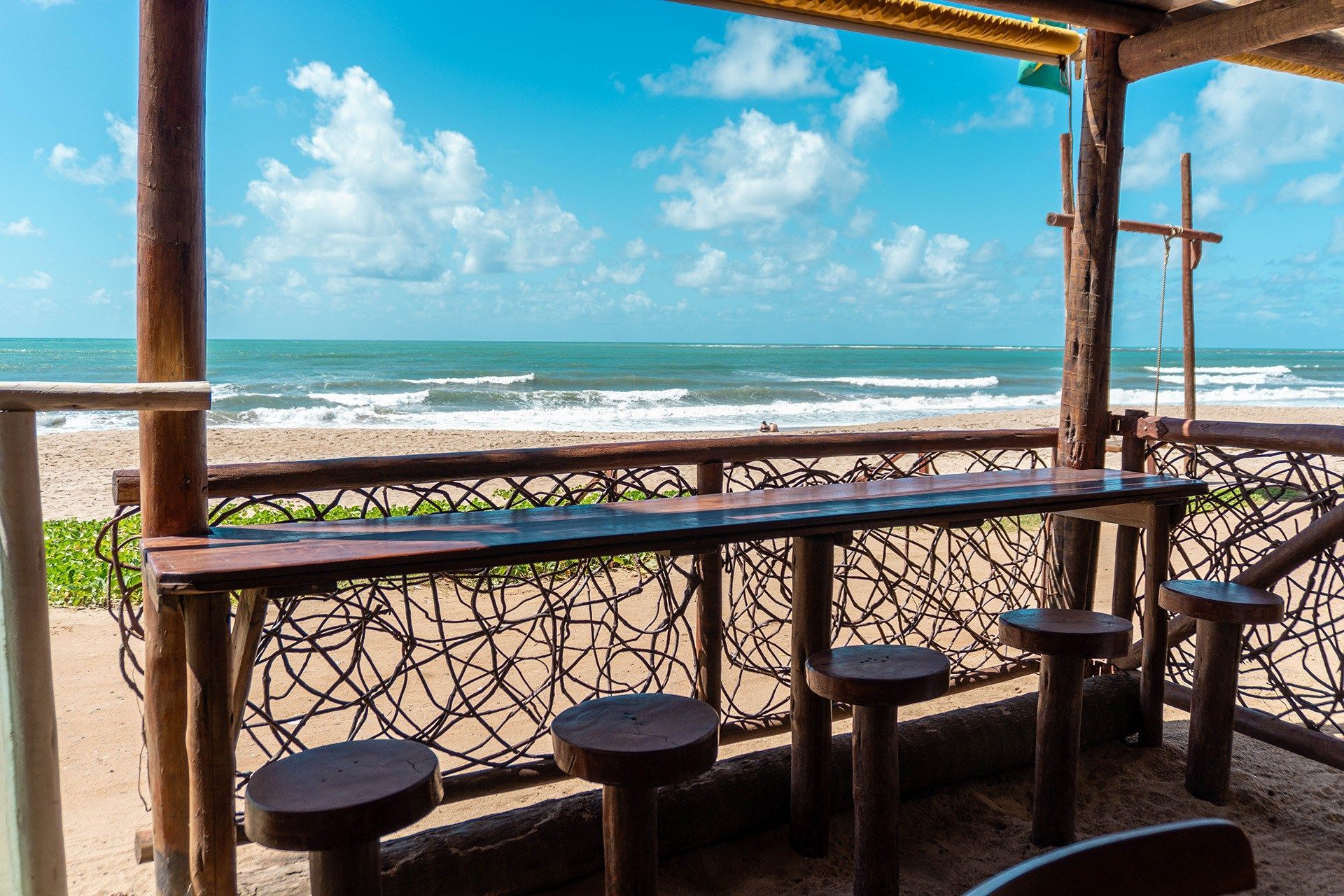 Bar e bancos de madeira com vista para uma praia de areia e o oceano sob um céu azul.