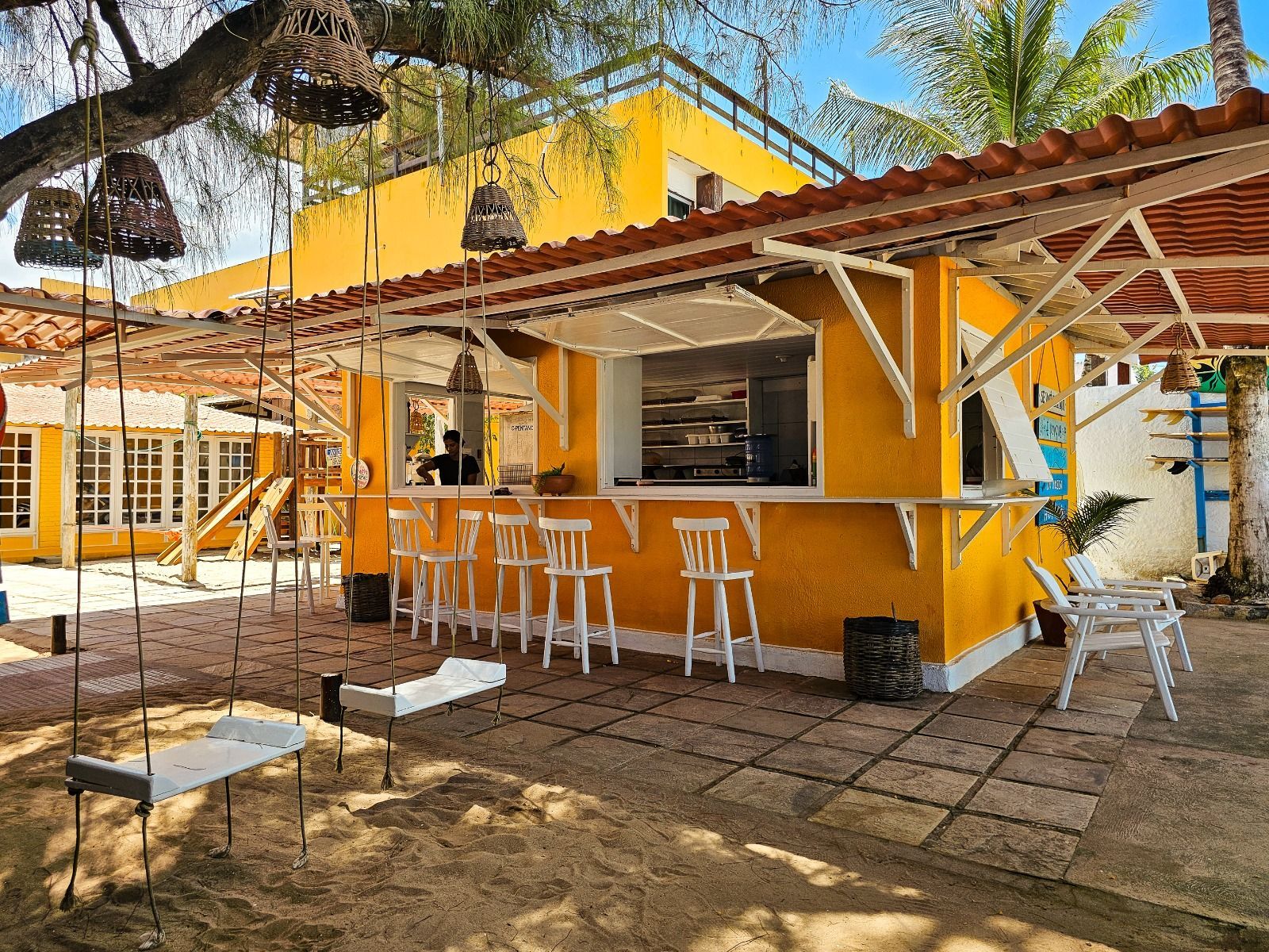 Quiosque de comida amarelo com balcão, decoração de praia, enfeites pendurados e uma árvore.