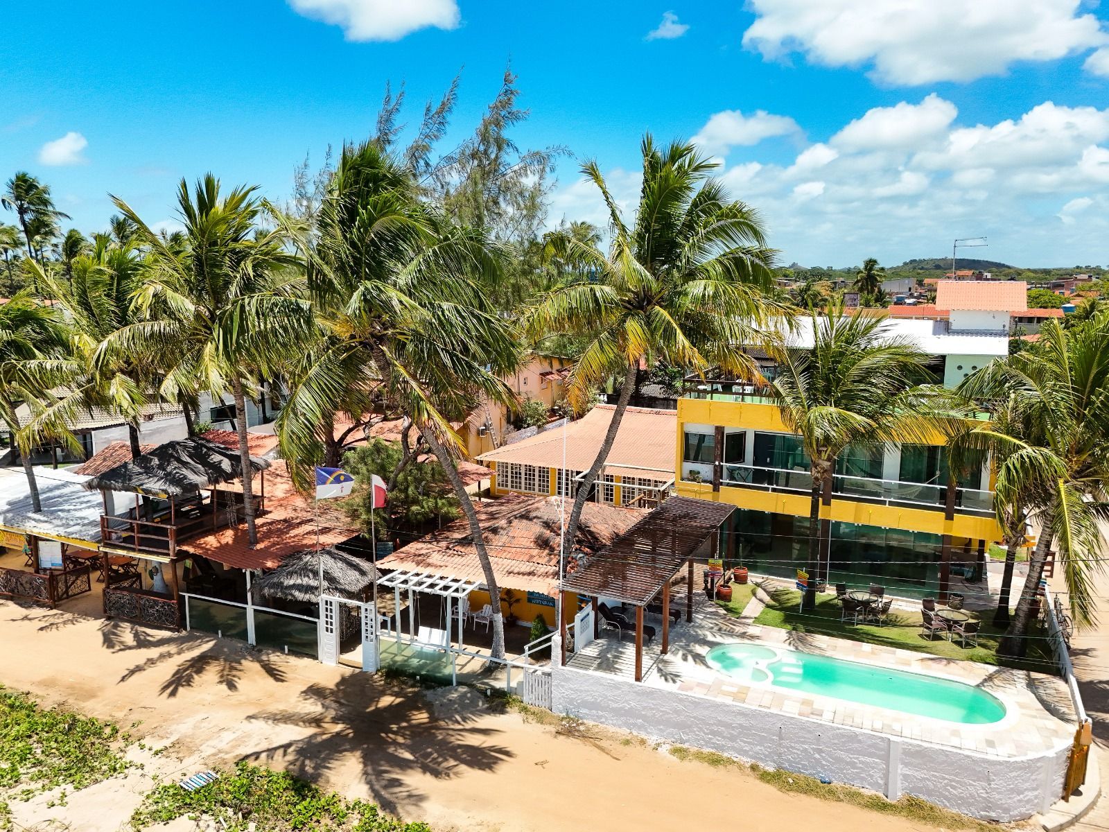Resort à beira-mar com piscina, palmeiras e edifícios amarelos e brancos sob um céu azul.