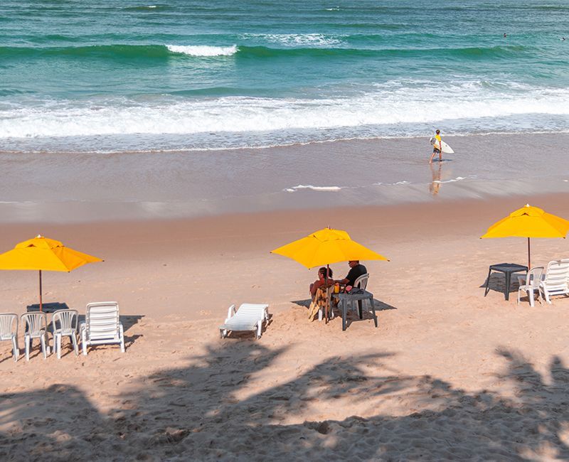 Cena de praia: guarda-sóis amarelos, cadeiras, pessoas e um surfista perto das ondas.