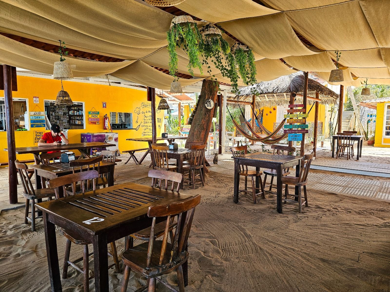 Restaurante ao ar livre com paredes amarelas, chão de areia, mesas e cadeiras de madeira e uma rede.