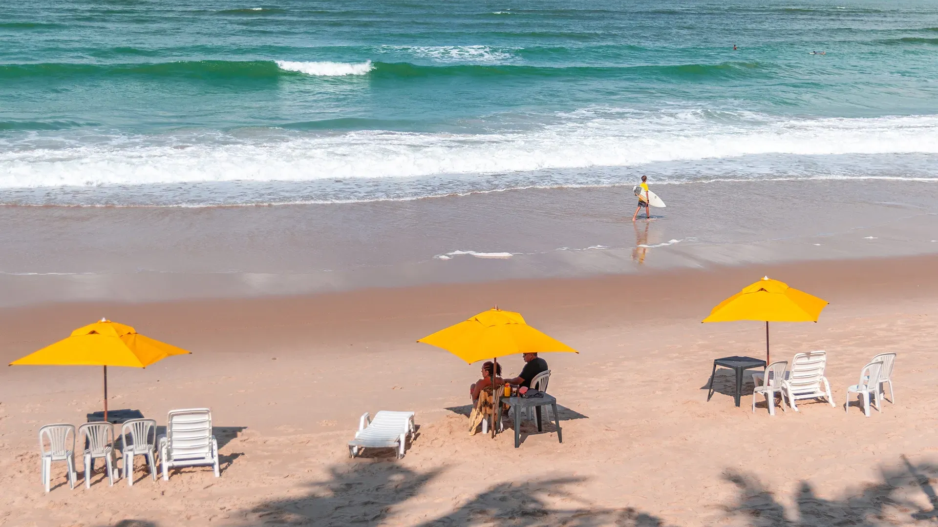 Praia de areia com mar turquesa. Guarda-sóis amarelos sombreiam mesas e cadeiras. Uma pessoa entra no mar com uma prancha de surfe.