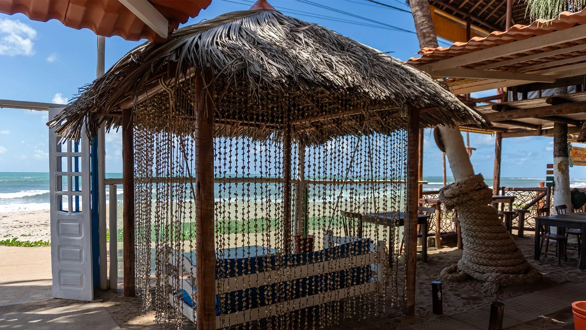 Gazebo com teto de palha em uma praia, viga de sustentação envolta em corda, areia branca e oceano ao fundo.