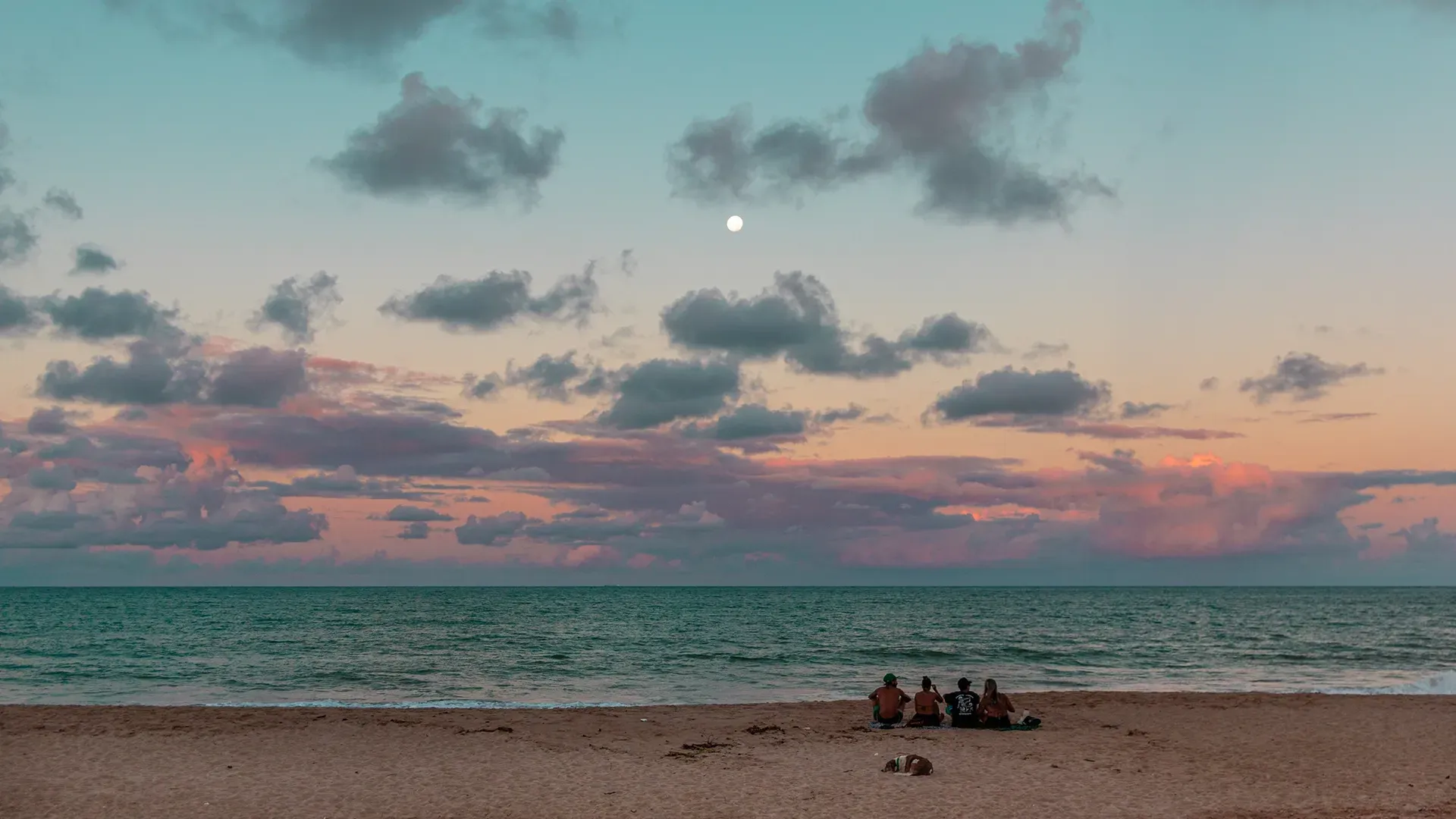 Pôr do sol na praia: Silhuetas sentadas na areia, observando o céu colorido com nuvens e a lua sobre o oceano.