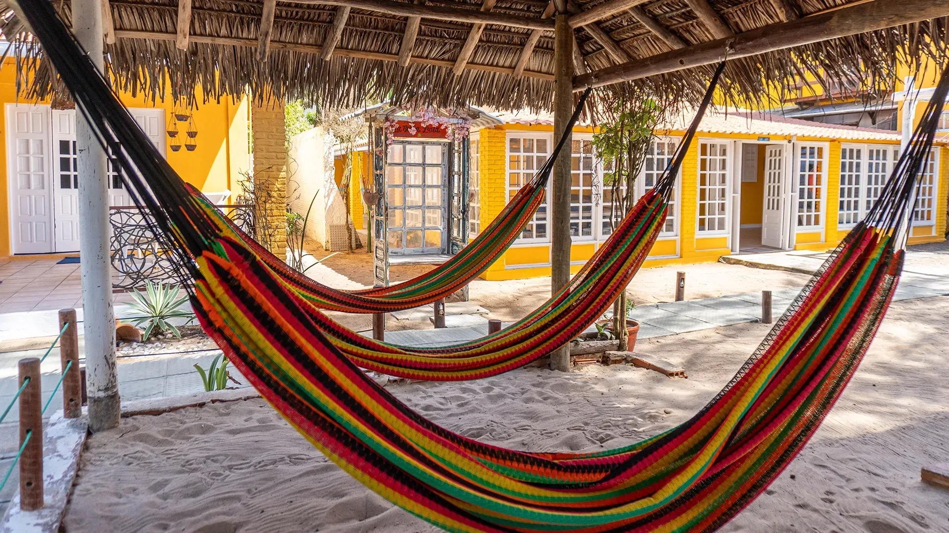 Redes penduradas sob um telhado de palha em um pátio de areia em frente a um prédio amarelo.