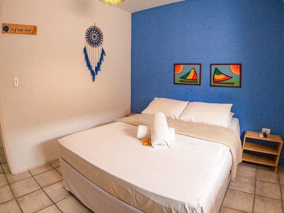 Quarto com cama branca, parede com detalhes em azul, obras de arte, filtro dos sonhos e pequena mesa de cabeceira de madeira.