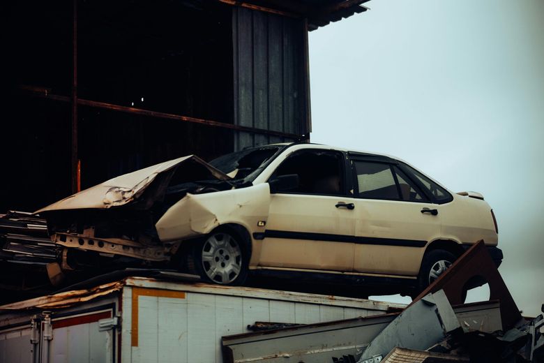 Damaged white car on top of a container, possibly in a junkyard, under a cloudy sky.