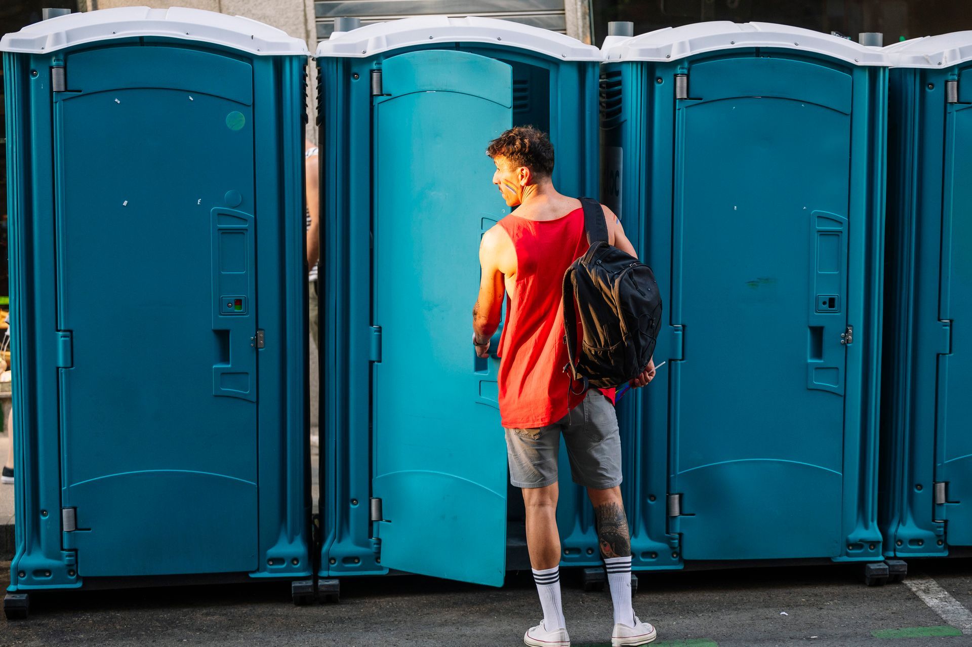 A man stands between two blue portable restrooms, one with the door open.
