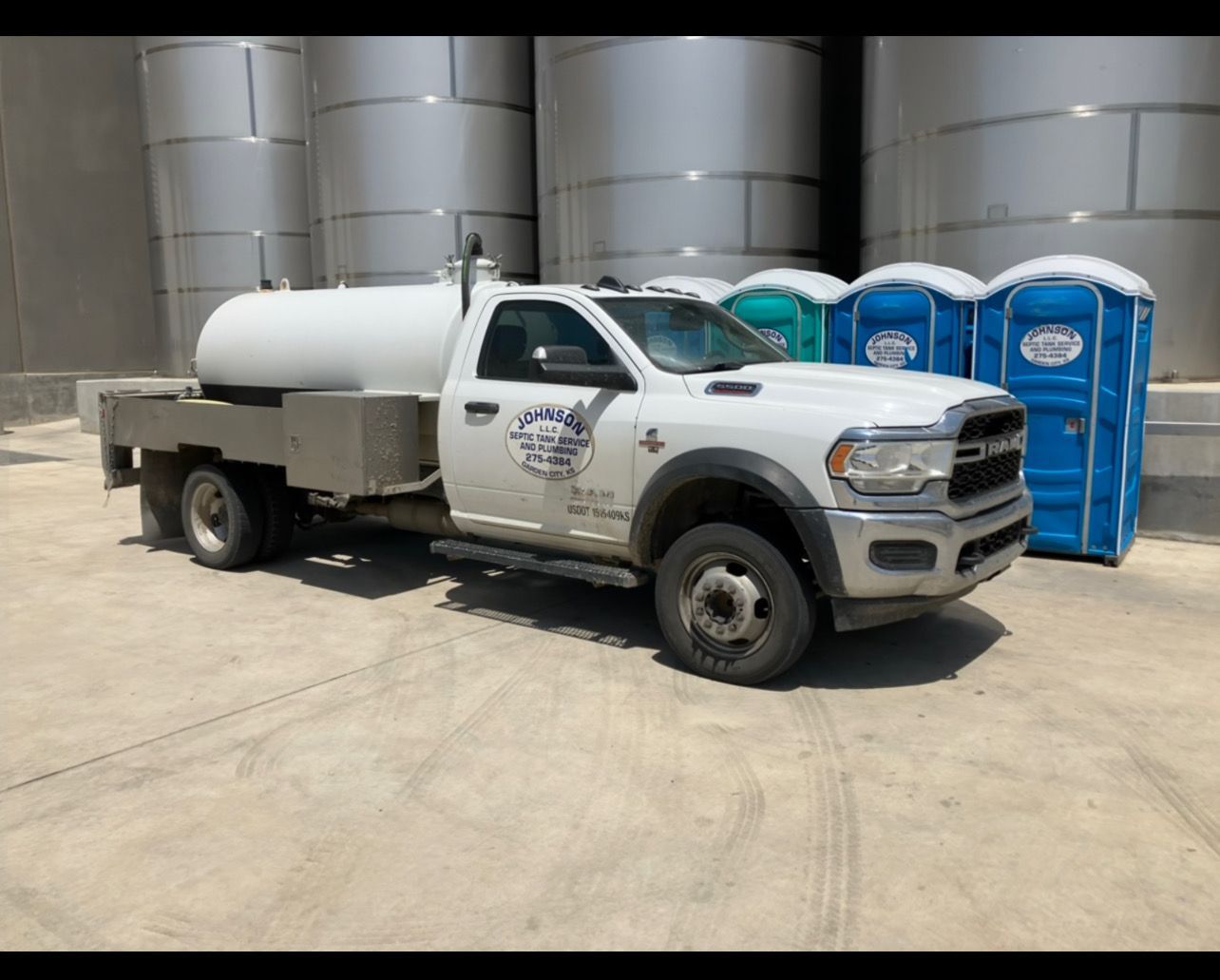 White truck with water tank parked next to portable toilets, in front of large metal tanks.