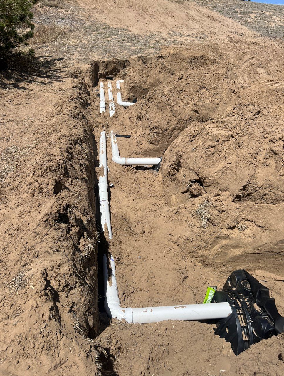 Trench with white PVC pipes laid within. Brown soil surrounds the pipes; setting is outdoors.