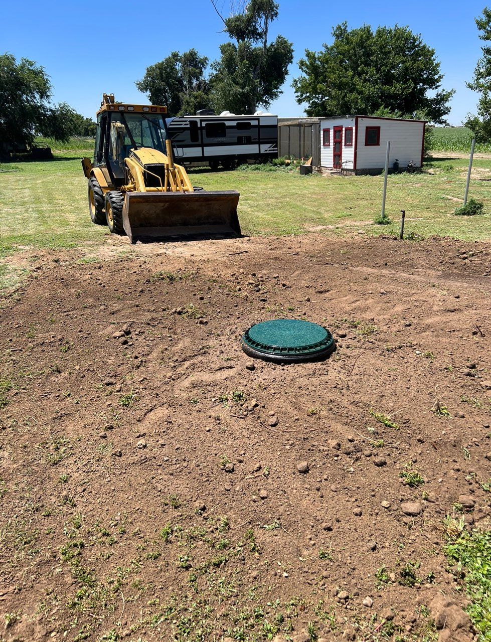 Backhoe near an open, dirt area with a septic tank lid; outdoor setting.