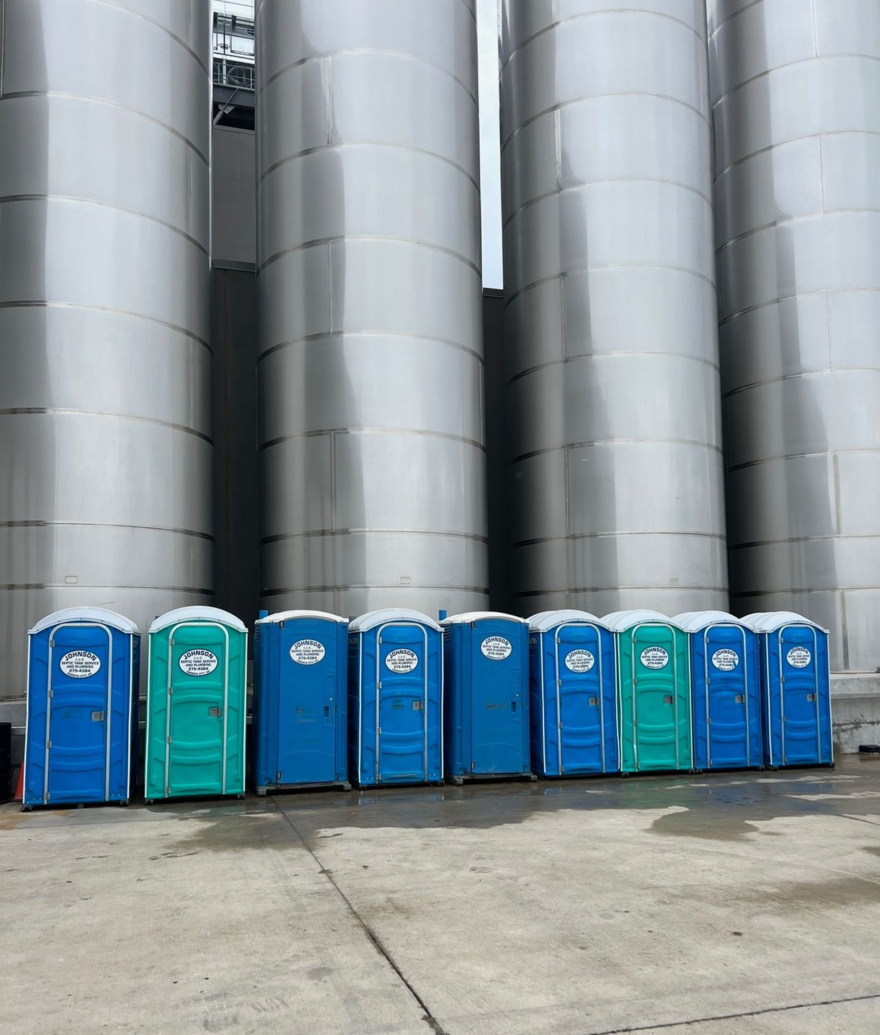 Portable toilets in various colors lined up in front of large metal silos.