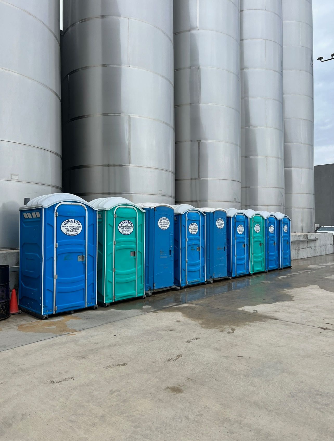 Row of blue and green portable toilets lined up near large metal industrial tanks.