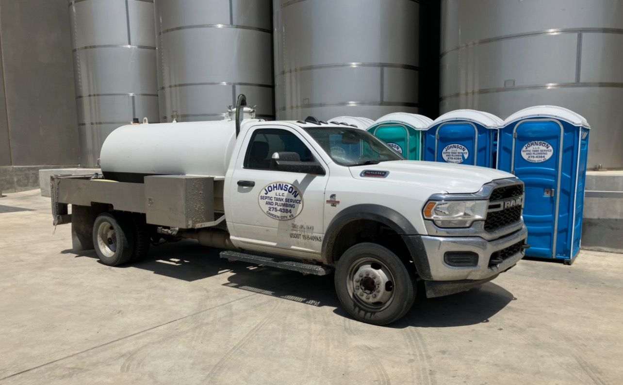 White truck with water tank, parked next to portable toilets, set in front of industrial tanks.