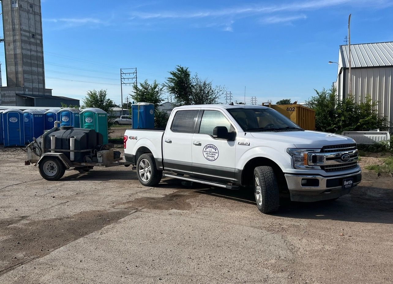 White truck towing a trailer with portable toilets. Blue sky, industrial setting.
