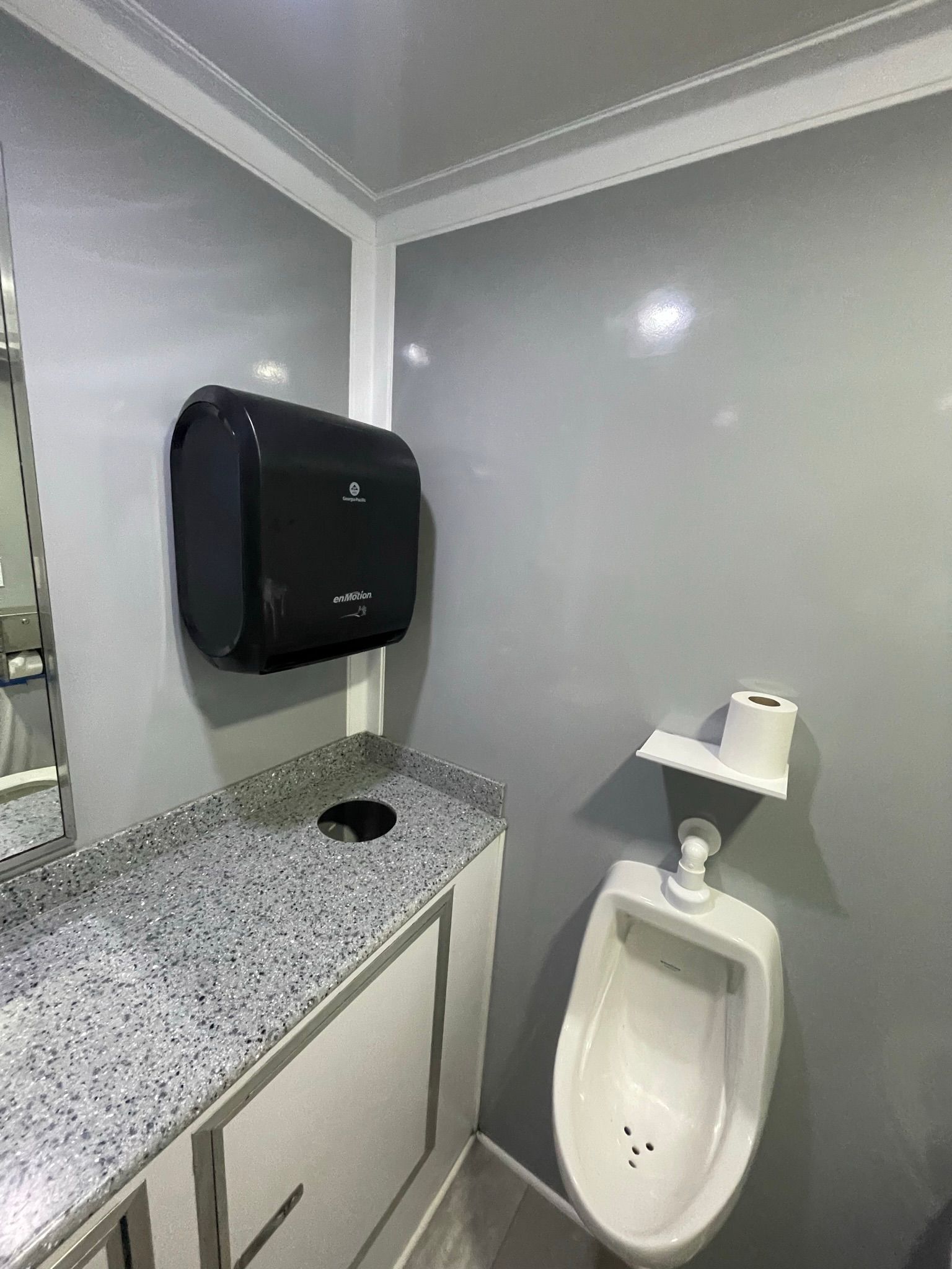 A portable restroom with a urinal, paper towel dispenser, and countertop with sink.