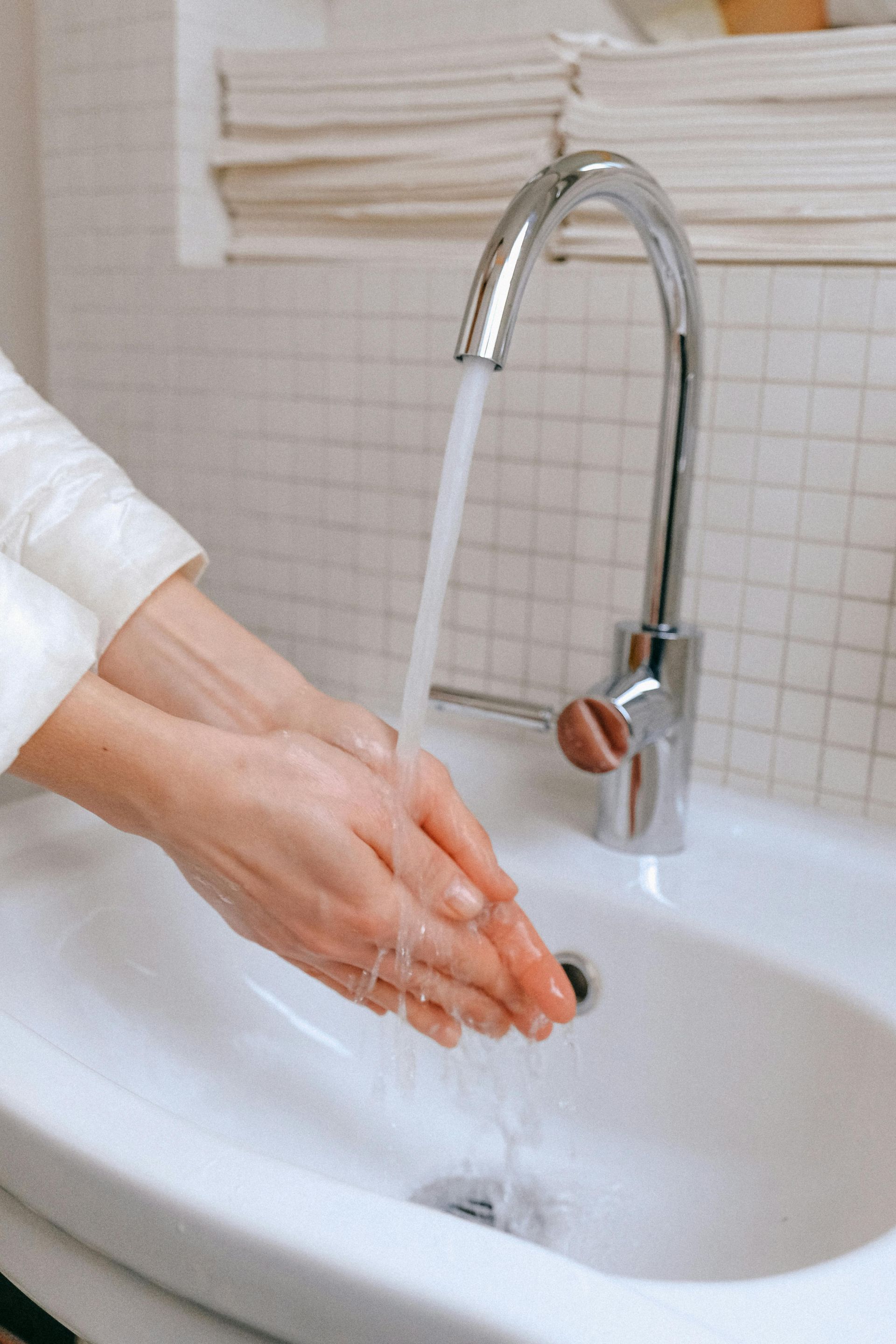 Hands are being washed under running water from a chrome faucet in a white-tiled bathroom.