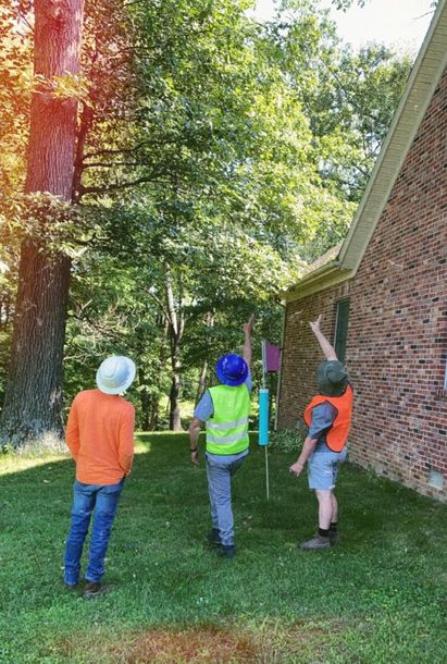 Three children in hard hats walking on grass beside a brick building and trees